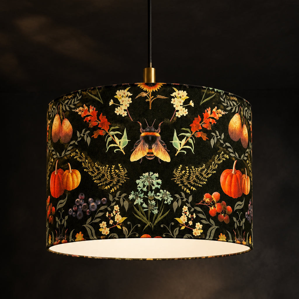 A drum pendant light shade with a black background and an intricate botanical and autumnal print featuring pumpkins, flowers, leaves, and a butterfly, suspended from a brass ceiling canopy against a dark textured wall.