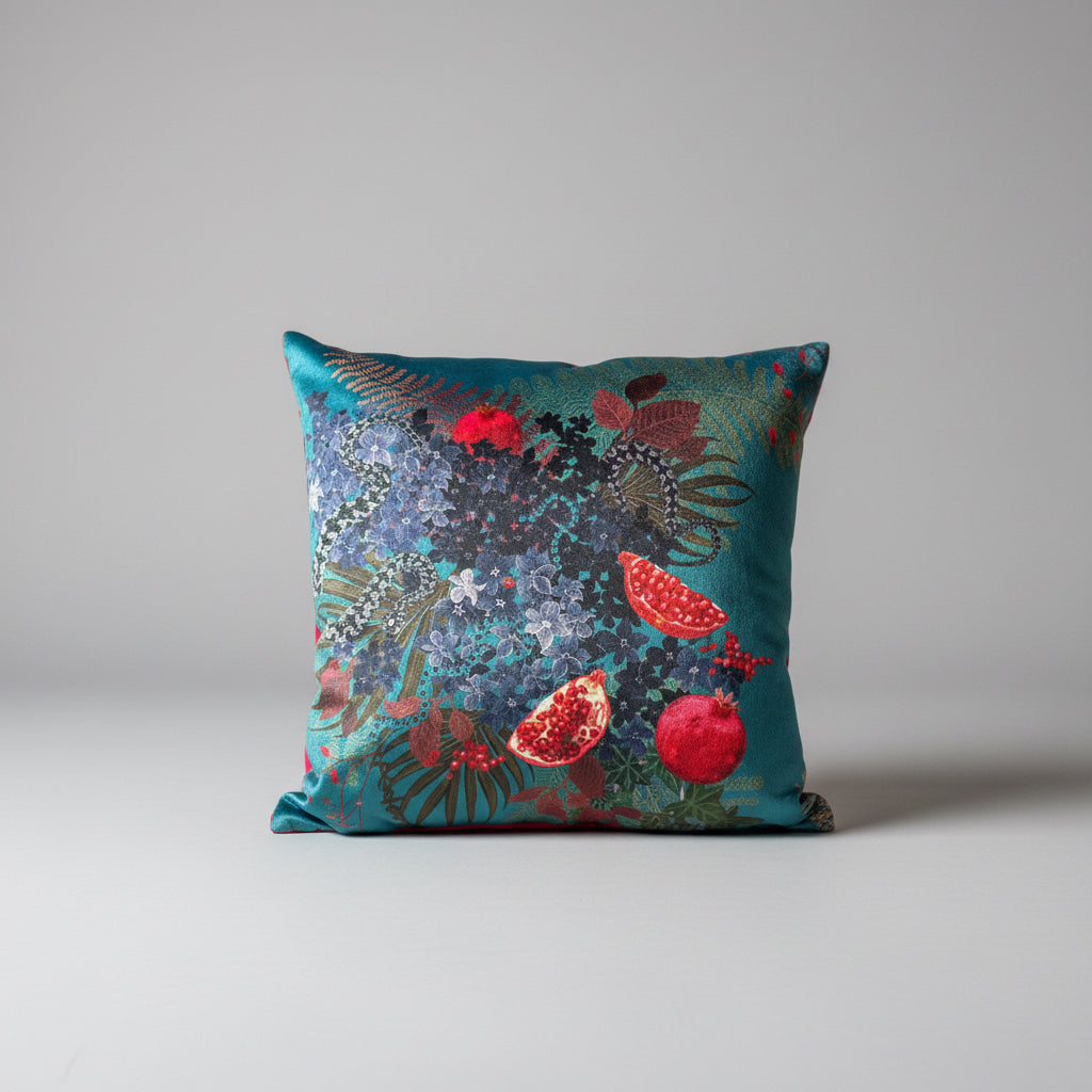 A luxury velvet cushion with a dramatic design featuring a ripe pomegranate, petals, an adder snake, and butterflies, displayed on an azure blue background. Becca Who