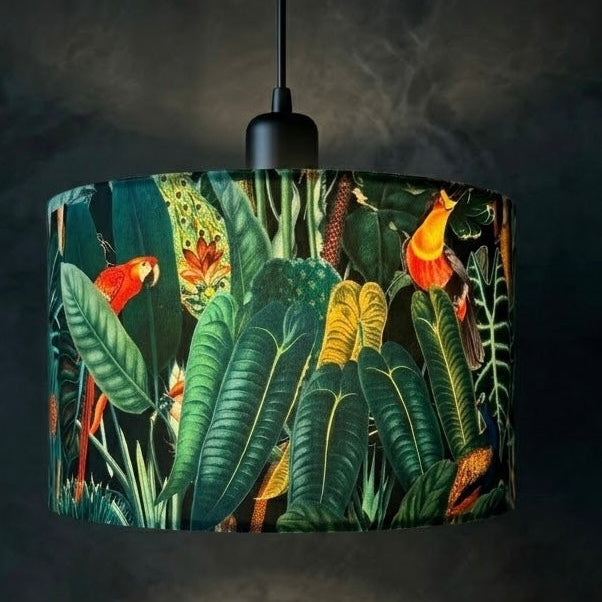 A hanging pendant lamp with a black ceiling canopy and cord, featuring a drum shade printed with a lush tropical jungle scene including large green leaves, red and orange birds, and yellow flowers, shown against a dark textured wall.
