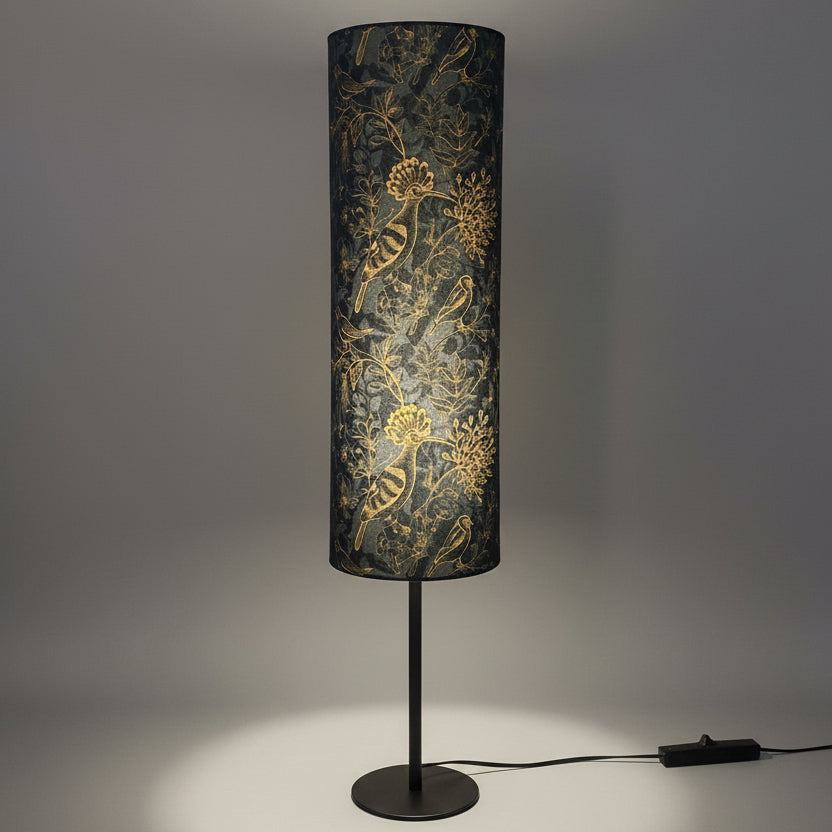 A tall cylindrical floor lamp with a dark grey and gold floral and bird  patterned shade, mounted on a slim black metal stand with a round base, set on a wooden floor against a textured gray wall.