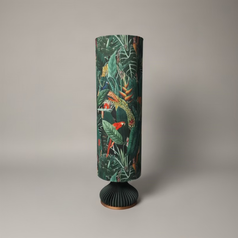 A tall cylindrical floor lamp with a dark green tropical jungle print featuring leaves, birds, and flowers, standing on a pleated dark green base over a wooden floor against a textured gray wall.