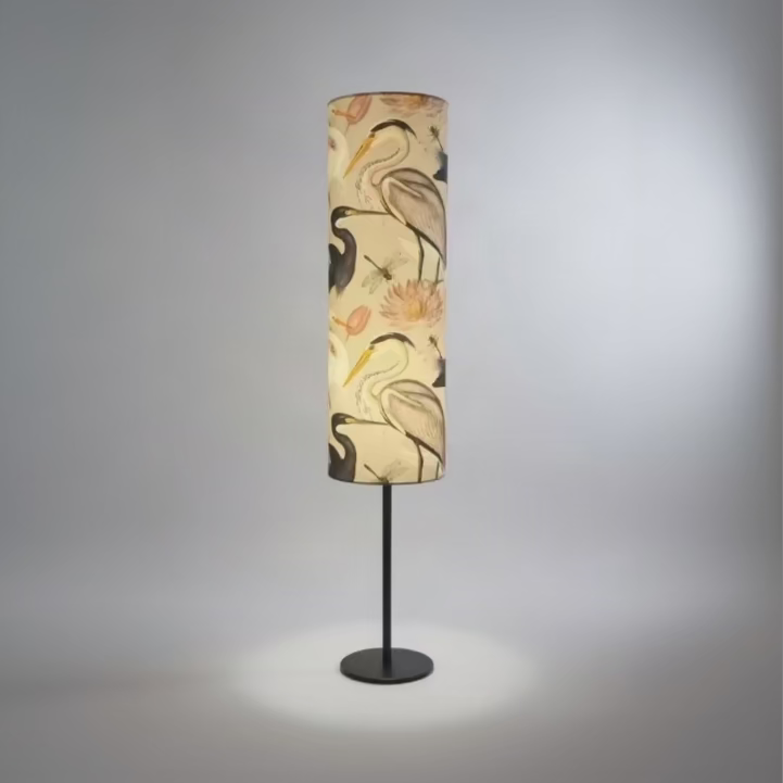 A floor lamp with a cylindrical shade featuring a printed pattern of herons, storks, and dragonflies on a cream background, illuminated from within and set on a dark metal stand.